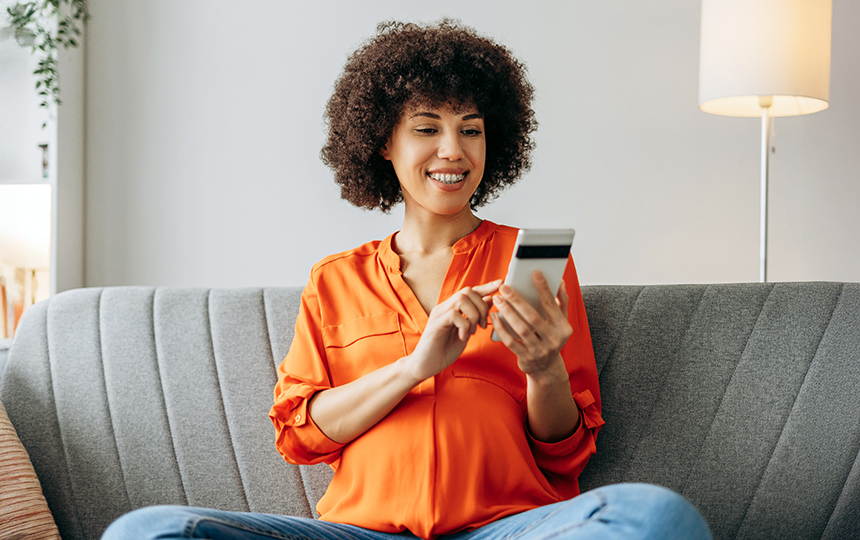 Mulher, vestindo camisa laranja, sentada no sofá usando o celular para realizar transações online, representando tendências em meios de pagamento digitais
