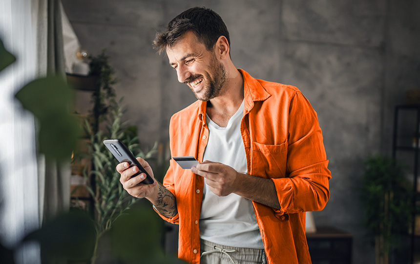 Homem sorridente usando camisa laranja segura um celular em uma mão e um cartão de crédito na outra, aparentando fazer uma compra online em um ambiente doméstico bem iluminado e decorado com plantas.