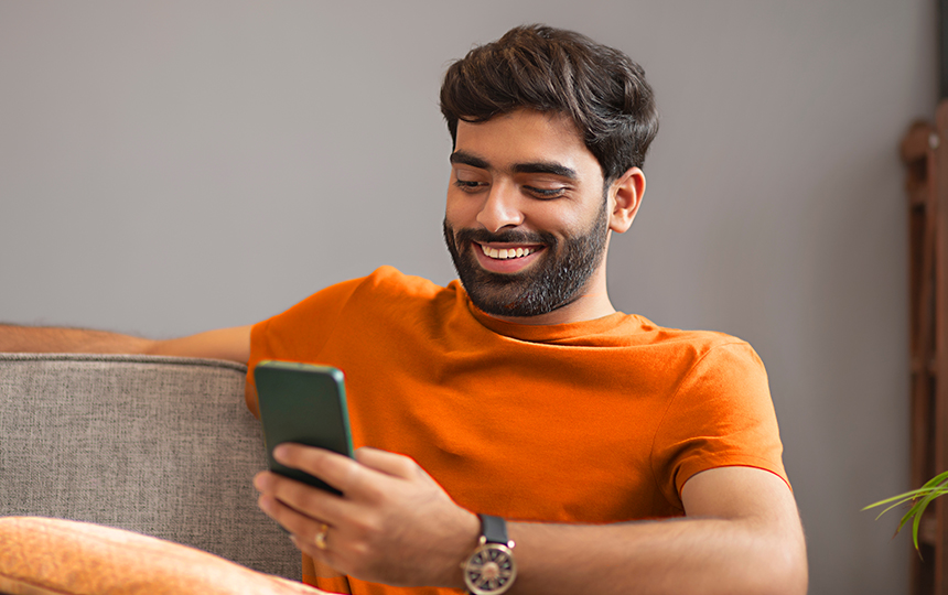 Homem sorridente, vestido com camisa laranha, relógio e aliança, está sentado, olhando para o celular em ambiente doméstico.