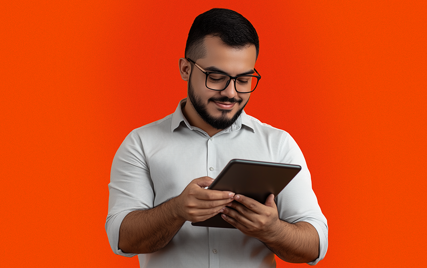 homem de óculos, camisa cinza e barba olhando um tablet e sorrindo imagem com fundo laranja 