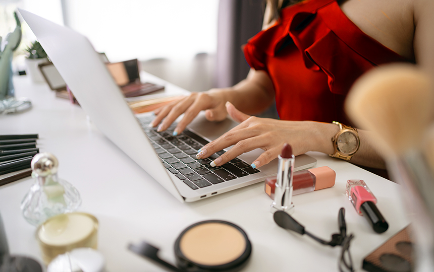 Mulher digitando em notebook sobre penteadeira com maquiagem, em ambiente iluminado e decorado, representando negócios digitais e e-commerce de beleza.