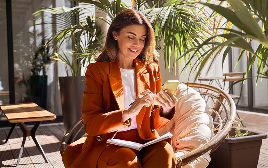 Mulher sorridente usando smartphone sentada em varanda com plantas, vestida de blazer laranja e caderno no colo, representando facilidade de realizar pagamentos digitais