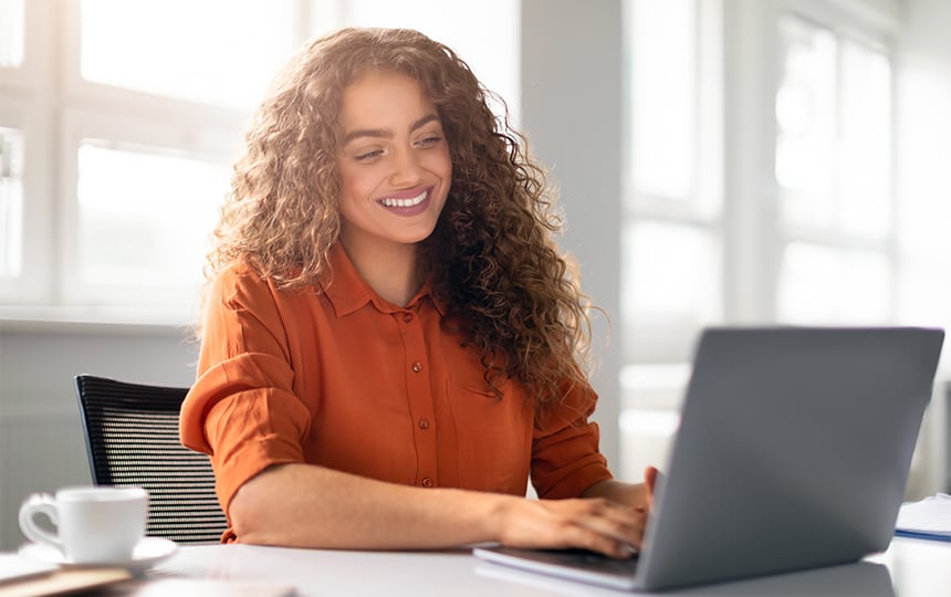 Mulher jovem sorridente com camisa terracota, trabalhando em um notebook sobre a mesa com xícara ao lado.